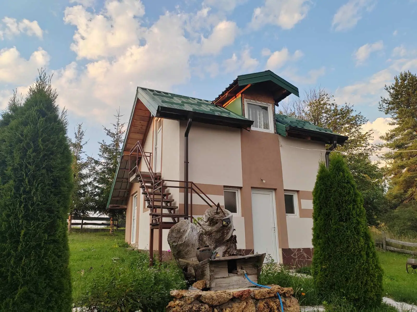 Vikendica kod kruške — zidana kućica sa zelenim limenim krovom, spoljnim stepeništem i česmom od panja